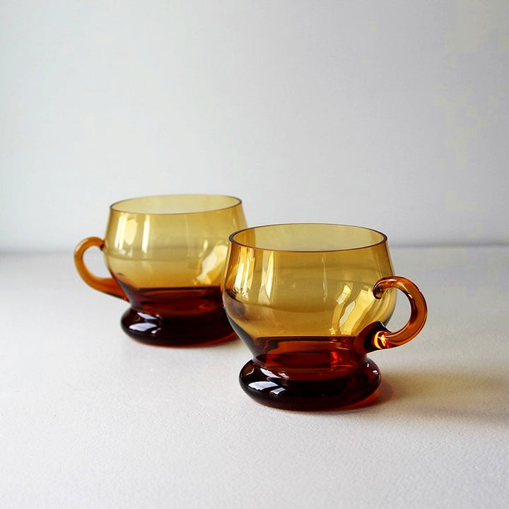 Zwei Vintage-Bowletassen aus bernsteinfarbenem Glas mit Standfuß und Henkel aus den 1970er Jahren.