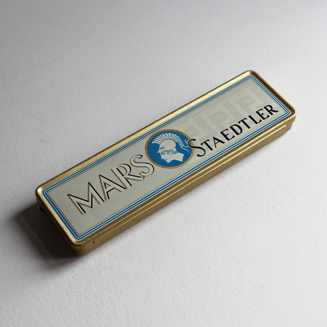 Alte längliche Mars Staedtler Blechdose mit cremeweißem Deckel, blauem Mars-Kopf-Logo, geometrischem Dekor und goldfarbenem Rand.