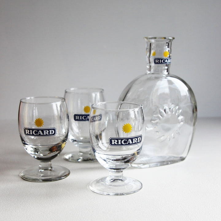 Vintage Ricard Pastis-Set aus einer klaren Glaskaraffe mit reliefartigem Sonnenmotiv und drei passenden Stielgläsern mit blau-gelbem Ricard-Logo.