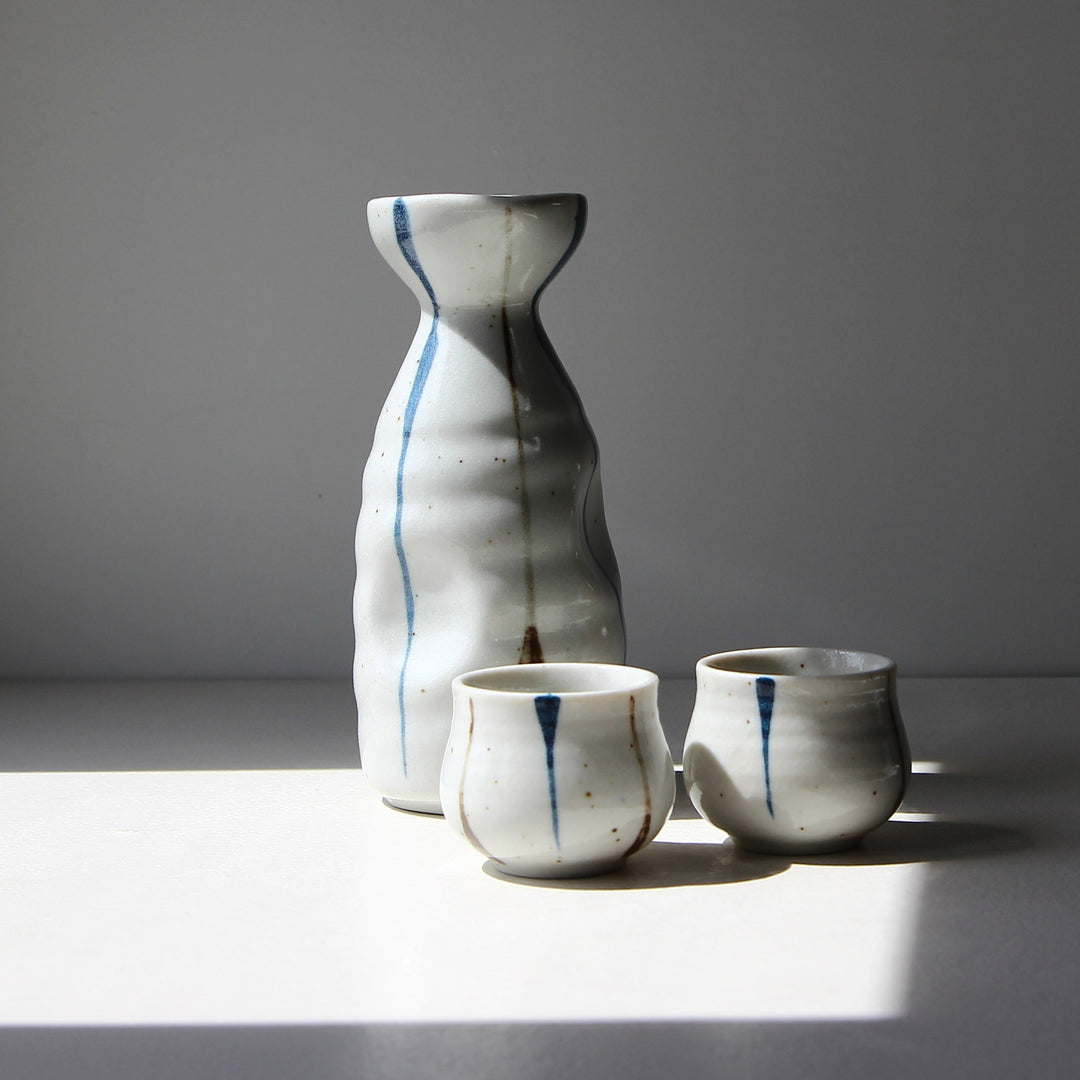 Japanisches Saké-Set Ligne Verticale aus Keramik mit Tokkuri-Karaffe und zwei Ochoko-Tassen in weißer Glasur mit blauen vertikalen Linien.