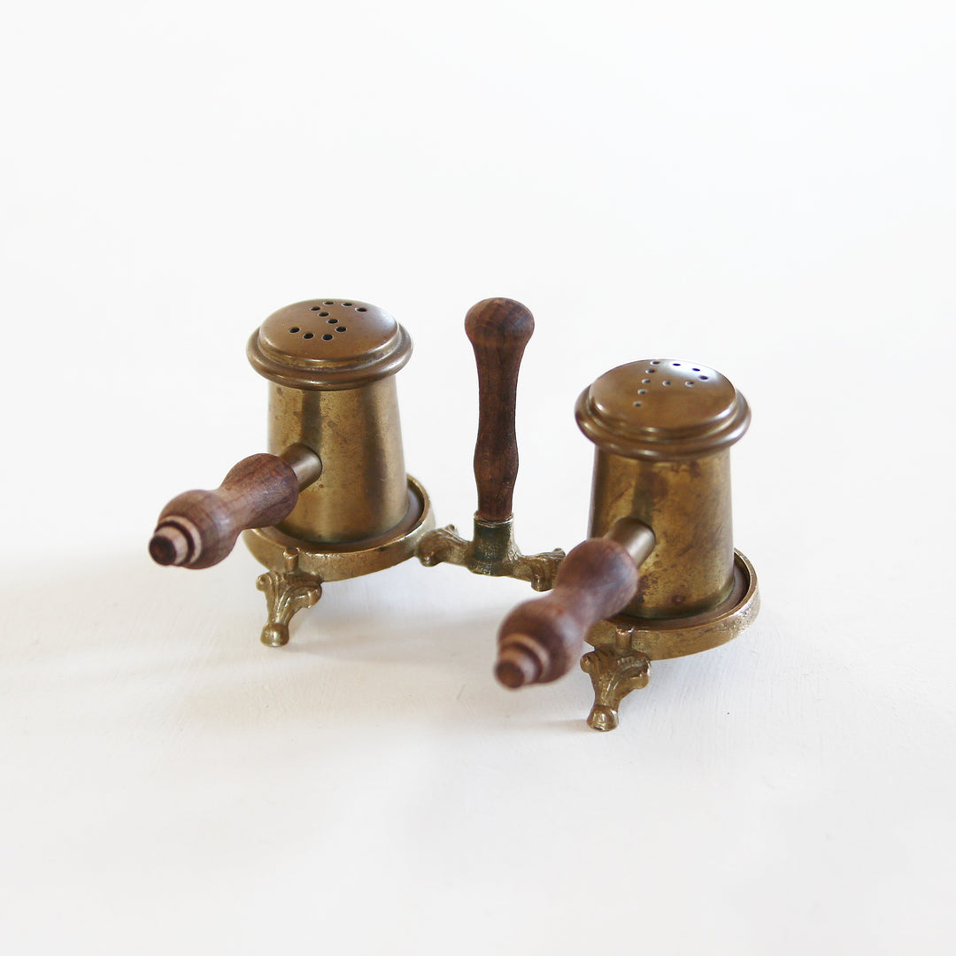 Altes Salz- und Pfefferstreuer-Set aus Messing mit zwei zylindrischen Streuern, gedrechselten Holzgriffen und gemeinsamer Halterung auf kleinen Füßen.