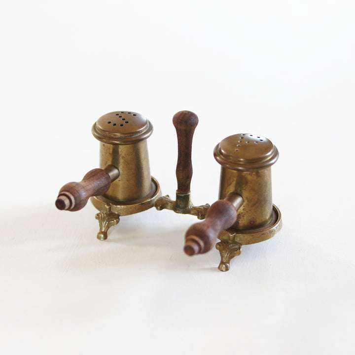 Altes Salz- und Pfefferstreuer-Set aus Messing mit zwei zylindrischen Streuern, gedrechselten Holzgriffen und gemeinsamer Halterung auf kleinen Füßen.