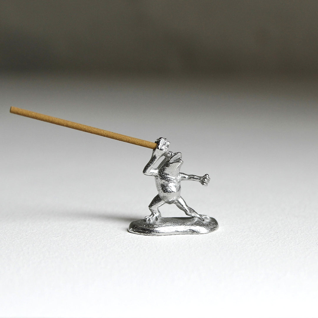 Pewter frog incense holder inspired by the Japanese Chōjū-giga scrolls, holding an incense stick. Crafted by Sumitani Saburo Shoten.