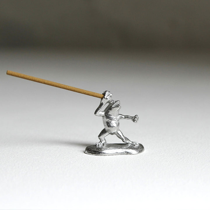 Pewter frog incense holder inspired by the Japanese Chōjū-giga scrolls, holding an incense stick. Crafted by Sumitani Saburo Shoten.